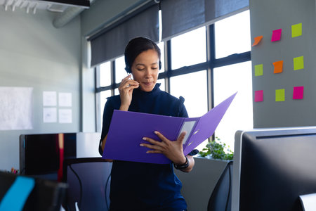 Asian woman office worker holding binder and speaking into headset microphone in open-plan office. Collaboration, communication, productivity, modern, professional, workspace, businessの写真素材