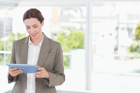Blazer-clad woman holding blue tablet near office glass wall with white window railing, copy space. Professional, corporate, modern, technology, workspace, communication, minimalisticの写真素材