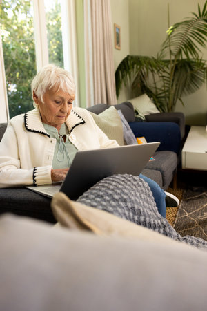 Senior woman using silver laptop while sitting on dark gray sofa in bright living room. Cozy,...