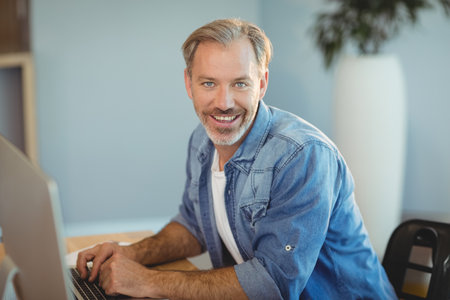Middle-aged man typing on computer keyboard at wooden desk in bright office with potted plant....