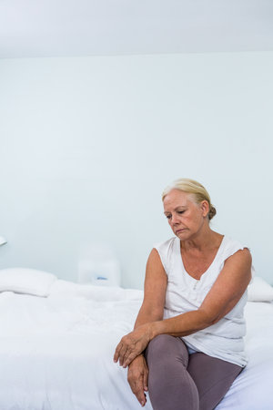 Woman is sitting on edge of white bed beside nightstand in white room, conveying somber mood. Minimalist, introspective, solitude, subdued, contemplative, tranquil, monochromeの写真素材