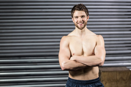 Shirtless athletic man standing with arms crossed in warehouse-style gym by wooden plyometric box. Strength, fitness, determination, industrial, training, male physique, motivationの写真素材