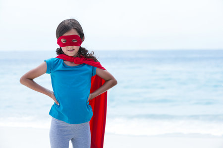 Girl standing on sandy shoreline wearing red superhero cape and eye mask, copy space. Adventure, resilience, empowerment, childhood, outdoor, playful, vibrantの写真素材