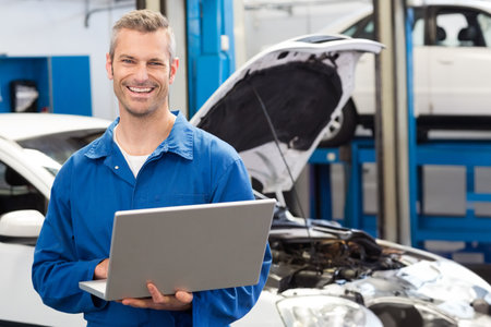 Male mechanic wearing blue coveralls using laptop inspecting lifted car with raised hood in garage....