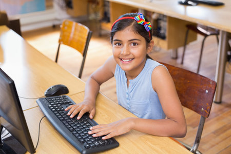 Indian child wearing rainbow bow headband typing on keyboard at computer lab desk with world map. Classroom, technology, education, youth, creativity, learning, innovationの写真素材