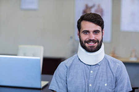 Male patient using silver laptop at medical clinic desk wearing cervical collar near anatomy charts. Rehabilitation, healthcare, physiotherapy, consultation, clinical, ergonomic, documentaryの写真素材