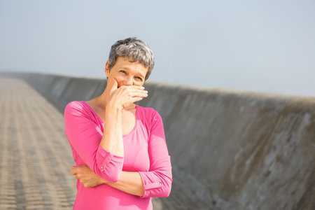 Senior woman standing on sloped coastal promenade wearing bright pink top and covering mouth. Scenic, outdoor, vibrant, leisure, coastal, tranquility, happinessの写真素材