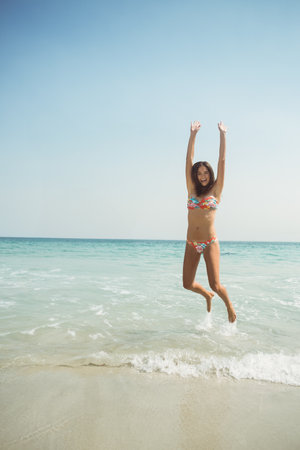 Woman jumping at sandy beach shoreline wearing floral-patterned bikini set with gentle ocean waves. Vacation, leisure, summer, outdoor, joyful, vibrant, landscapeの写真素材