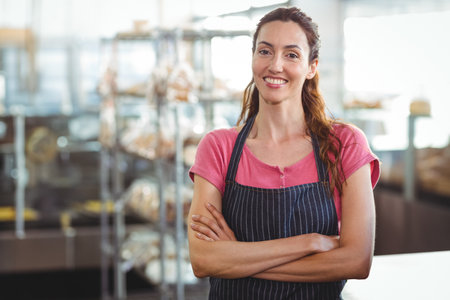 Female baker standing behind counter in bakery wearing striped apron and smiling with bread shelves....