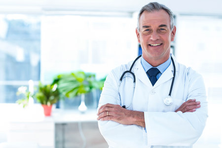 Mature adult male doctor standing arms crossed in coat by windows holding stethoscope, copy space. Healthcare, professionalism, wellness, consultation, modern, clean, brightの写真素材