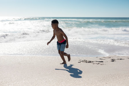 African American boy running along wet sand shoreline with ocean waves and striped swim trunks. Children, leisure, adventure, outdoor, summer, vitality, natureの写真素材