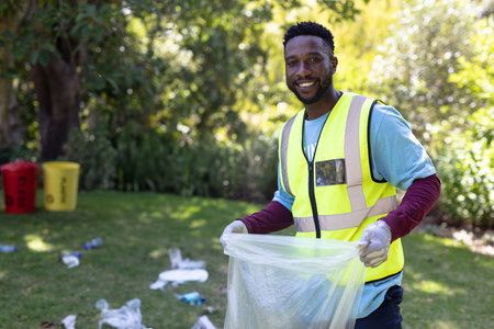 African American male volunteer picking litter on park lawn in vest holding trash bag, copy space....