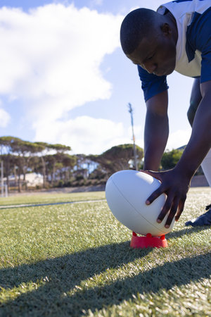 African American male athlete in jersey positioning rugby ball on kicking tee on pitch, copy space. Athlete, sports, outdoor, landscape, athleticism, competition, energyの写真素材