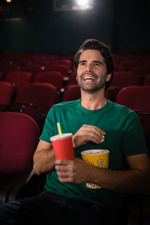 Popcorn tub and disposable cup sitting on red cushioned theater seats, under soft projector glow. Entertainment, leisure, theater, cinema, relaxation, enjoyment, ambientの写真素材