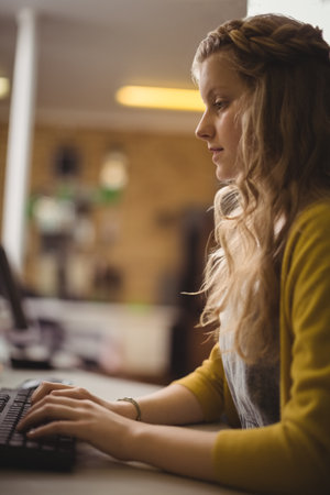 Female child wearing yellow cardigan and bracelet typing on keyboard at desk near bookshelves. Child, education, study, learning, cozy, workspace, homeofficeの写真素材