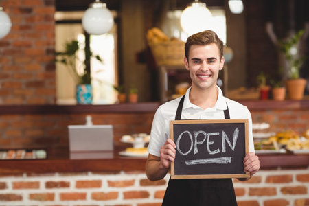 Male barista standing behind brick counter holding open chalkboard sign in bakery, copy space. Cafe,...