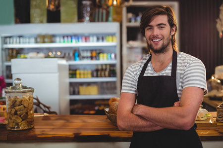 Male barista standing behind wooden cafe counter displaying pastries, jar, fridge, copy space. Bakery, hospitality, vintage, inviting, artisanal, warm, boutiqueの写真素材