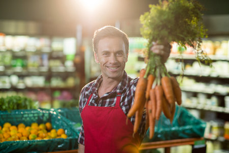 Male produce clerk smiling and presenting fresh carrots beside mesh orange bins at supermarket. Freshness, health, agriculture, market, eco, vibrant, nourishmentの写真素材