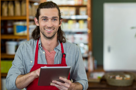 Male store clerk wearing red apron, holding digital tablet, checking inventory in shop aisle. Retail, shopping, technology, commerce, customer service, organization, presentationの写真素材