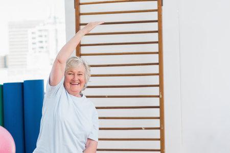 Senior woman stretching right arm overhead at fitness room by stall bars, mats and stability ball. Wellness, flexibility, rehabilitation, seniorhealth, exerciseequipment, mindfulness, activeagingの写真素材