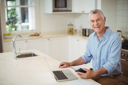 Senior man sitting at bright modern home kitchen holding smartphone and using laptop, copy space. Contemporary, tech, lifestyle, interior, relaxation, warmth, connectivityの写真素材