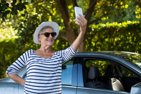 Senior woman wearing sun hat on driveway holding smartphone and taking selfie by silver sedan. Retirement, leisure, vacation, outdoor, lifestyle, tranquility, eleganceの写真素材