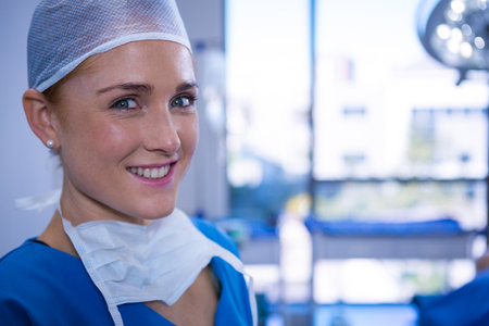 Female surgeon standing in operating room wearing surgical cap and mask under surgical light. Medical, healthcare, professional, hospital, clinic, hygiene, brightの写真素材