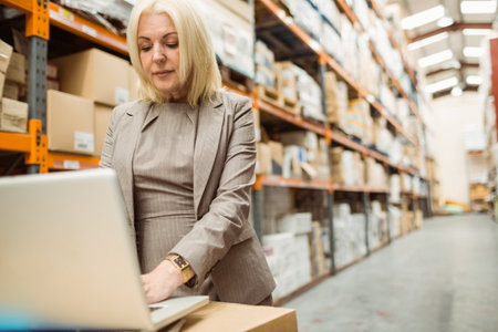 Woman in suit checking inventory in warehouse aisle using laptop beside boxes on racks, copy space. Industrial, logistics, efficiency, workspace, technology, organization, professionalの写真素材