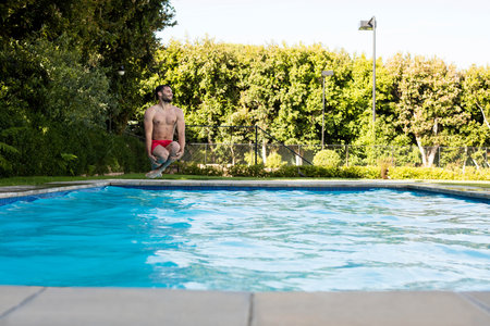 Fit man in red trunks crouching at concrete pool edge preparing to dive into blue water. Athletic, recreation, leisure, summer, outdoor, lifestyle, wellnessの写真素材