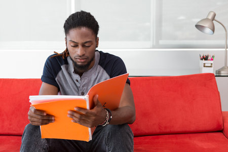 African American man sitting on red sofa reading orange binder in study with lamp, pencil holder....