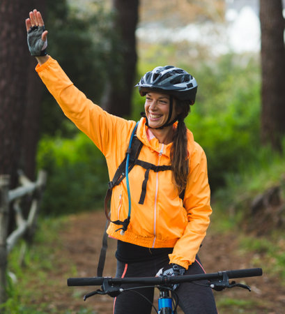 White female cyclist wearing helmet and hydration pack waving on forest dirt trail by wooden fence. Adventure, outdoor, fitness, exploration, nature, activity, vitalityの写真素材