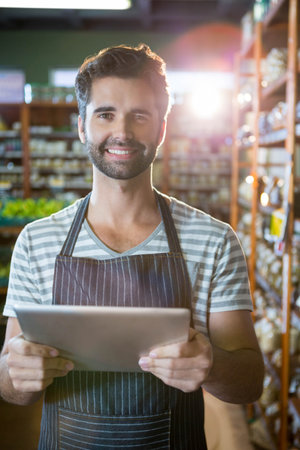 Adult male clerk wearing apron, holding tablet, checking stock in grocery aisle with wooden shelves....
