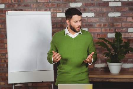 Man wearing sweater presenting in office with brick wall at flip chart beside plant and laptop....
