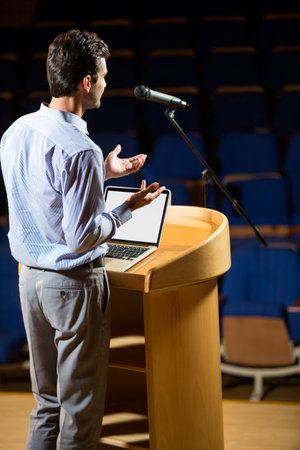 Speaker gesturing behind wooden podium with laptop and microphone on stage amid empty blue chairs. Public speaking, presentation, education, conference, professional, leadership, speechの写真素材