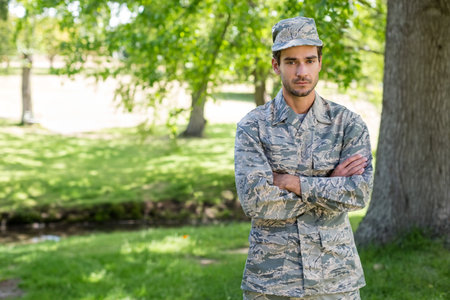 Male soldier crossing arms in park in camouflage uniform and cap by tree, stream, copy space. Military, outdoor, natural, rugged, resilience, strength, camouflageの写真素材