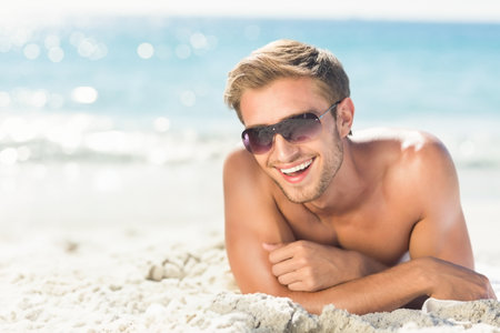 Shirtless man lying on white sand at beach by ocean wearing tinted sunglasses, smiling under sun. Beach, leisure, relaxation, fashion, outdoor, serenity, lifestyleの写真素材