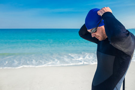 Man adjusting blue swim cap and securing tinted goggles on white sandy beach facing turquoise sea. Adventure, sport, athletic, aquatic, wellness, activity, seasideの写真素材