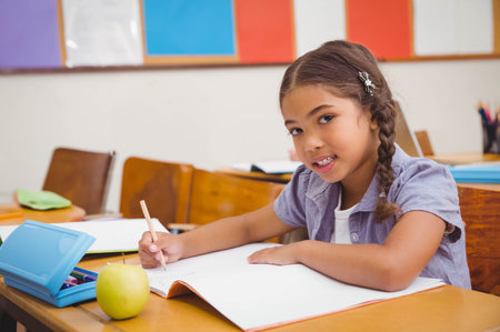 Child student writing in notebook at school desk with green apple, blue pencil case, copy space. Education, childhood, learning, classroom, stationary, studying, school suppliesの写真素材