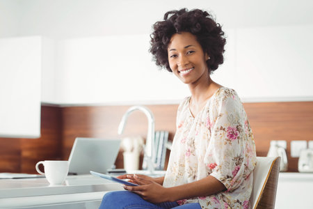 African American woman holding tablet next to coffee mug and laptop on kitchen counter, copy space. Modern, minimalist, cozy, lifestyle, technology, domestic, casualの写真素材