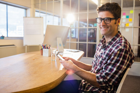 Mid-adult man sitting in office holding smartphone amid monitor pen holder sticky notes, copy space. Professional, collaboration, modernity, workplace, communication, tech-savvy, productivityの写真素材