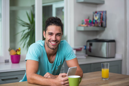 Man in his twenties sitting at kitchen counter in t shirt holding smartphone beside coffee mug. Contemporary, lifestyle, minimalistic, domestic, casual, vibrant, cozyの写真素材