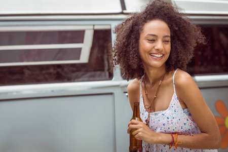 African American woman leaning on blue camper van outside holding brown glass bottle, copy space. Bohemian, nostalgic, vibrant, adventure, leisure, outdoor, travelの写真素材