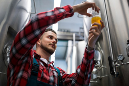 Man standing in brewery room holding clear beer sample glass near pressure gauge and steel tanks....