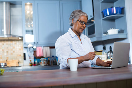 Senior African American woman checking credit card at kitchen counter using laptop with white mug. Technology, senior, domestic, lifestyle, modern, wellness, casualの写真素材