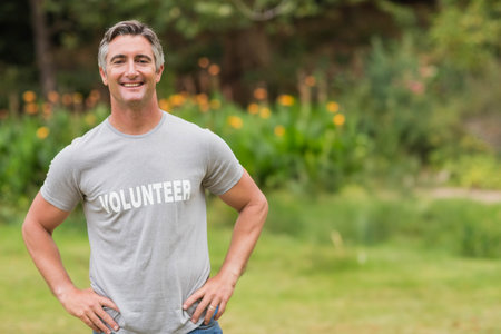 Middle-aged man standing with hands on hips in garden by flower bed wearing gray volunteer T-shirt. Nature, greenery, outdoor, community, environmental, cheerful, casualの写真素材