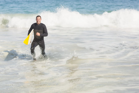 Middle aged man in black wetsuit emerging from ocean at beach carrying white and yellow surfboard....
