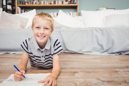 School-age boy wearing striped polo drawing with blue marker on paper in living room. Youth,...