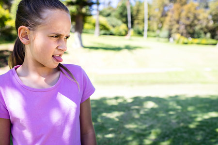 Female child grimacing with tongue out wearing purple tee, looking left in sunny park, copy space....