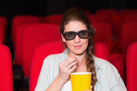 Teenage white female wearing black 3D glasses holding yellow popcorn bucket in darkened auditorium. Cinema, entertainment, leisure, vibrant, enjoyment, immersive, casualの写真素材