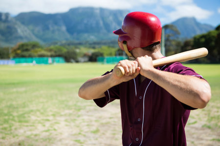 Baseball player holding wooden bat and standing in batter box wearing red helmet on sunlit field. Athlete, sports, outdoor, active, recreation, competition, athleticismの写真素材